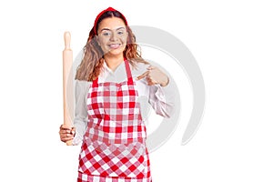Young latin woman wearing apron holding kneader pointing finger to one self smiling happy and proud