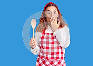 Young latin woman wearing apron holding wooden spoon covering mouth with hand, shocked and afraid for mistake