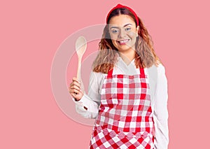 Young latin woman wearing apron holding wooden spoon looking positive and happy standing and smiling with a confident smile