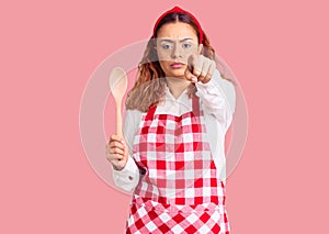 Young latin woman wearing apron holding wooden spoon pointing with finger to the camera and to you, confident gesture looking