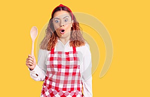 Young latin woman wearing apron holding wooden spoon scared and amazed with open mouth for surprise, disbelief face