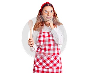 Young latin woman wearing apron holding wooden spoon serious face thinking about question with hand on chin, thoughtful about