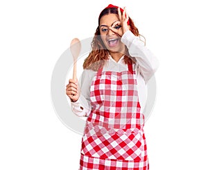 Young latin woman wearing apron holding wooden spoon smiling happy doing ok sign with hand on eye looking through fingers