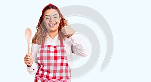 Young latin woman wearing apron holding wooden spoon smiling happy and positive, thumb up doing excellent and approval sign