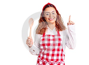 Young latin woman wearing apron holding wooden spoon surprised with an idea or question pointing finger with happy face, number