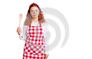 Young latin woman wearing apron holding wooden spoon thinking attitude and sober expression looking self confident