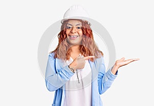 Young latin woman wearing architect hardhat amazed and smiling to the camera while presenting with hand and pointing with finger