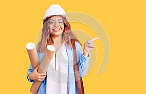 Young latin woman wearing architect hardhat holding blueprints smiling happy pointing with hand and finger to the side