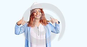 Young latin woman wearing architect hardhat looking confident with smile on face, pointing oneself with fingers proud and happy