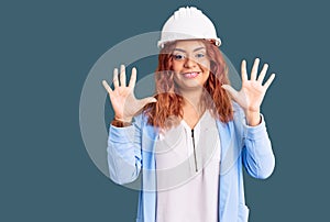 Young latin woman wearing architect hardhat showing and pointing up with fingers number ten while smiling confident and happy