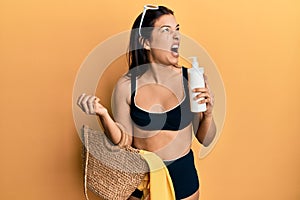 Young latin woman wearing bikini and hat holding summer wicker handbag and sunscreen angry and mad screaming frustrated and
