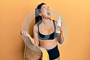 Young latin woman wearing bikini and hat holding summer wicker handbag and sunscreen angry and mad screaming frustrated and