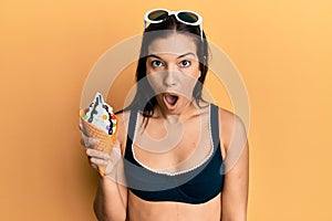 Young latin woman wearing bikini holding ice cream scared and amazed with open mouth for surprise, disbelief face