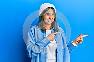 Young latin woman wearing cute wool cap pointing aside worried and nervous with both hands, concerned and surprised expression