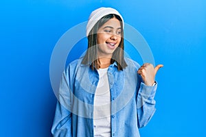 Young latin woman wearing cute wool cap smiling with happy face looking and pointing to the side with thumb up