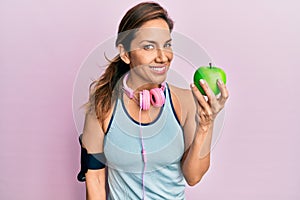 Young latin woman wearing gym clothes, using headphones and eating green apple looking positive and happy standing and smiling