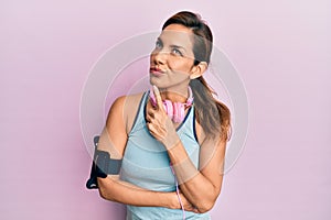 Young latin woman wearing gym clothes and using headphones thinking concentrated about doubt with finger on chin and looking up
