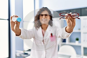 Young latin woman wearing optician uniform holding optometrist glasses and contact lenses at optical clinic