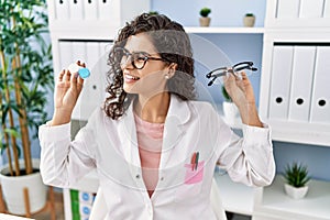 Young latin woman wearing optician uniform holding optometrist glasses and contact lenses at optical clinic