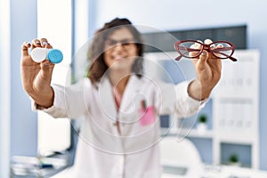 Young latin woman wearing optician uniform holding optometrist glasses and contact lenses at optical clinic