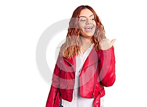 Young latin woman wearing red leather jacket smiling with happy face looking and pointing to the side with thumb up