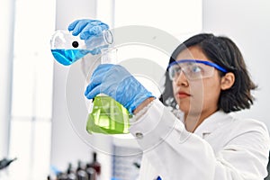 Young latin woman wearing scientist uniform pouring liquid on test tube at laboratory