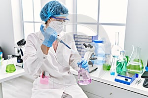 Young latin woman wearing scientist uniform pouring liquid on test tube at laboratory