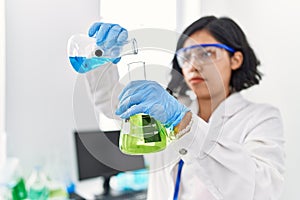 Young latin woman wearing scientist uniform pouring liquid on test tube at laboratory