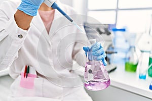 Young latin woman wearing scientist uniform pouring liquid on test tube at laboratory