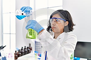 Young latin woman wearing scientist uniform pouring liquid on test tube at laboratory