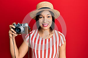 Young latin woman wearing summer hat holding vintage camera looking positive and happy standing and smiling with a confident smile