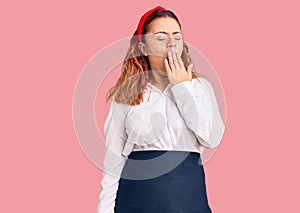 Young latin woman wearing waitress apron bored yawning tired covering mouth with hand