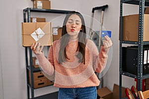 Young latin woman working at small business ecommerce holding credit card looking at the camera blowing a kiss being lovely and