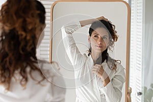 Young latina female in bathrobe looking at mirror creating hairdo