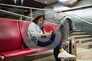 Young latina sitting in the airport terminal with her suitcase looking at the mobile phone. Latina chat with her cell phone while