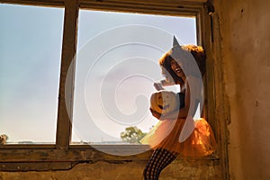 Young Latina woman dressed as a witch and sitting in a window frame laughing loudly and reaching into a Halloween pumpkin
