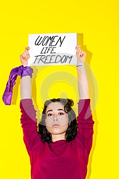Young Latina woman with a purple scarf on her wrist holding a sign reading WOMEN LIFE FREEDOM.