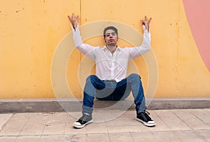 Young latino guy sitting and leaning against a yellow wall with his arms open up. Modern urban clothing