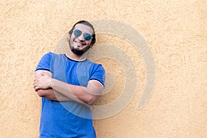 Young latino man with glasses, crossed arms and long hair, smiling looking at the camera, leaning against a yellow wall