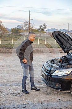 Young Latino man looking at the engine of his broken down car on the side of the road