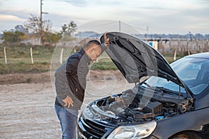 Young Latino man looking at the engine of his broken down car on the side of the road