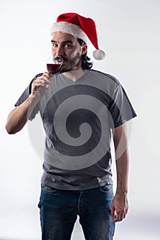Young Latino man in Santa Claus hat, drinking a glass of wine and looking at camera.