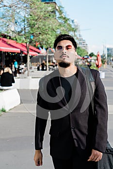 Young Latino man walking confidently down a street on a sunny day with a bag slung over his shoulder and wearing sunglasses. Trust