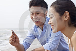 Young laughing couple looking at seashell at the waters edge, China