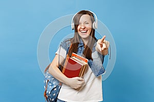 Young laughing joyful woman student with backpack headphones listening music hold school book pointing index finger on