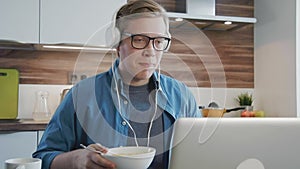 Young laughing man watching streaming movie on laptop at home eating cereals with headphones