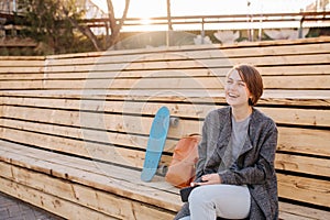 Young laughing woman with a skateboard is sitting on a 2-stage bench.