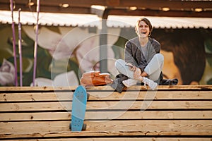 Young laughing woman with a skateboard is sitting on a 2-stage bench.