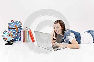 Young laughing woman student in denim clothes working on laptop pc computer lying near globe, backpack, school books