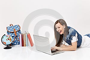 Young laughing woman student in denim clothes working on laptop pc computer lying near globe, backpack, school books